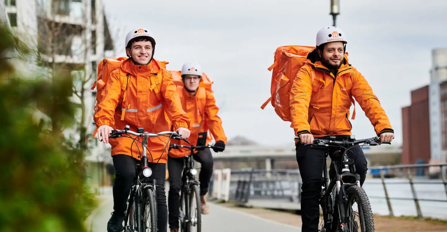 Drie maaltijdbezorgers op de fiets in oranje regenjassen met bezorgtas